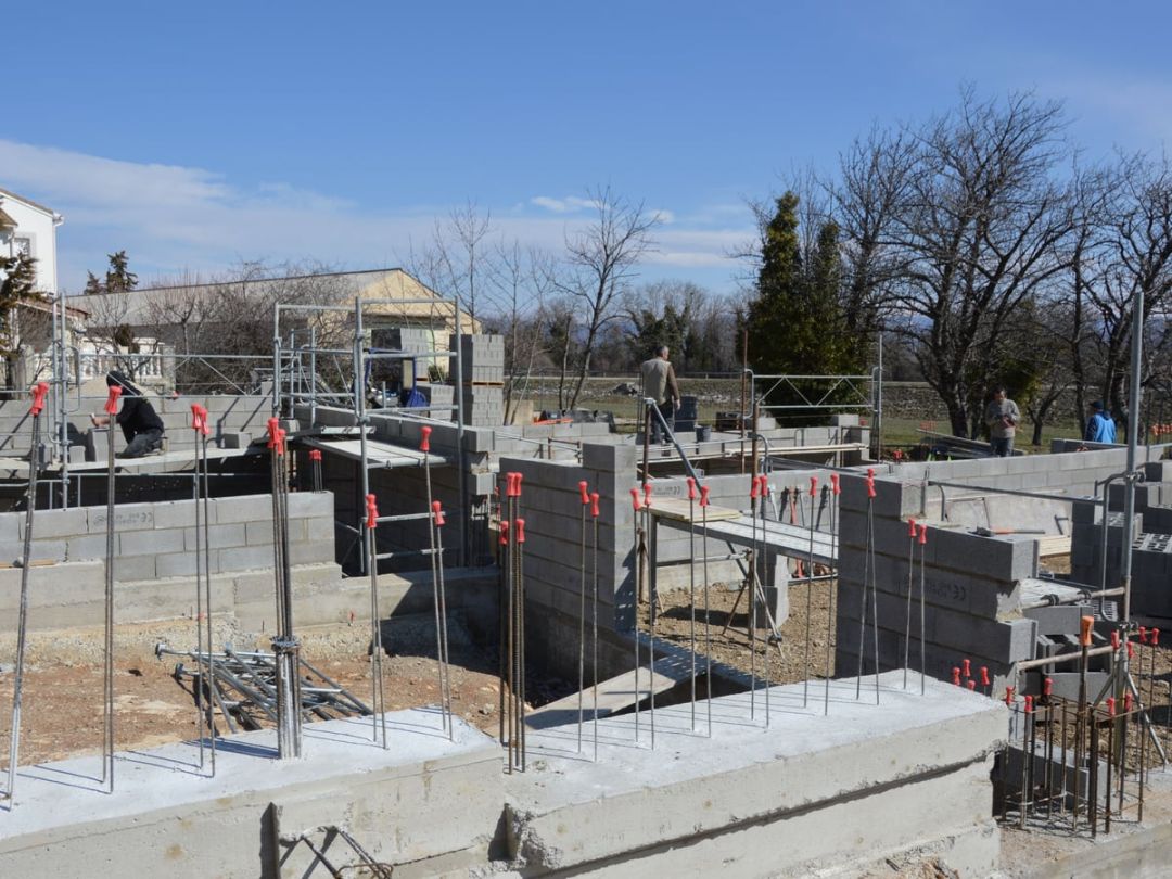Baustelle mit betonierten Fundamenten, Schalung und Bewehrung, im Hintergrund arbeiten zwei Personen