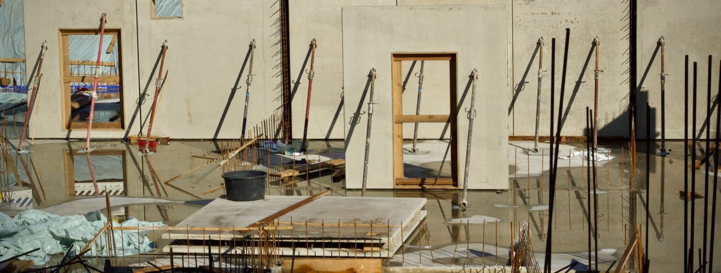 Rohbau eines Wohngebäudes: gestellte Betonwände mit Fenster und Türöffnungen, abgestützt mit Schrägstützen, davor Bewehrungsstahl